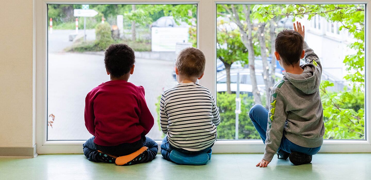 Das Bild zeigt drei Kinder, die mit dem Rücken zur Kamera vor einem großen Fenster sitzen und nach draußen schauen. 